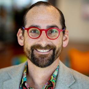 Headshot of Derek Laney — smiling, red round glasses, grey blazer over a patterned shirt.